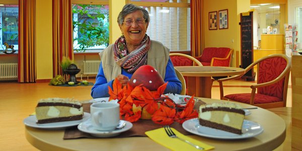 Eine ältere Dame sitzt am gedeckten Kaffeetisch und lacht in die Kamera 